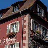 Ein rotes Gebäude mit weißen Fensterläden, Blumenkästen und einem verzierten Schmiedeeisenschild unter einem steilen Ziegeldach vor blauem Himmel.