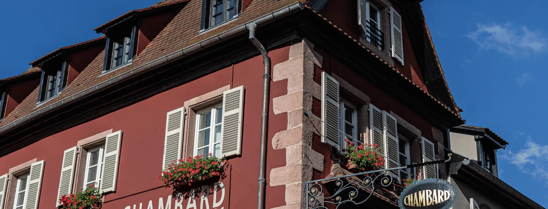 Ein rotes Gebäude mit weißen Fensterläden, Blumenkästen und einem verzierten Schmiedeeisenschild unter einem steilen Ziegeldach vor blauem Himmel.