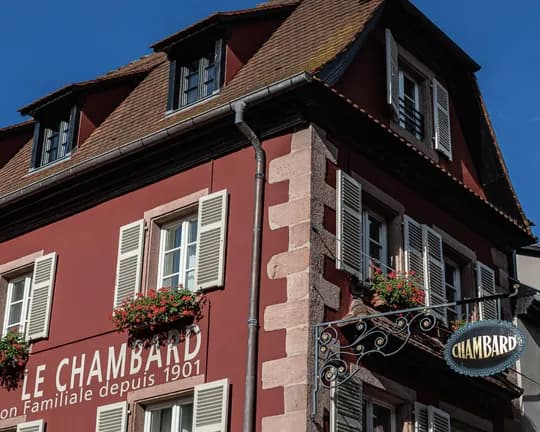 Red building with white shutters, flower boxes, and an ornate wrought-iron sign beneath a steep tile roof against a blue sky.