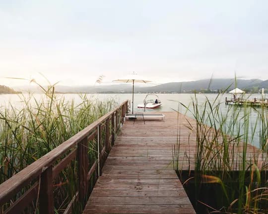 Wooden pier extends over a calm lake; tall reeds in the foreground, parasols and loungers on the deck, a small boat nearby, hills in the distance.