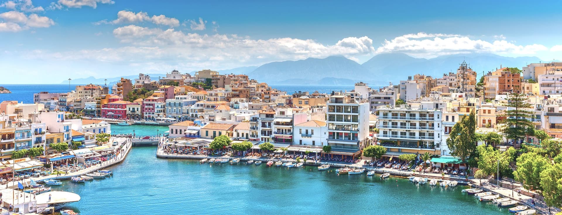Scenic view of Agios Nikolaos, Crete, with blue water, boats, and buildings under a sunny sky. Mountains are visible in the background.