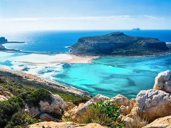 Θέα του μαγευτικού τοπίου της λιμνοθάλασσας του Μπάλου, στην Κρήτη, με τα τιρκουάζ νερά, τη λευκή άμμο και τους βραχώδεις βράχους κάτω από έναν καταγάλανο ουρανό.