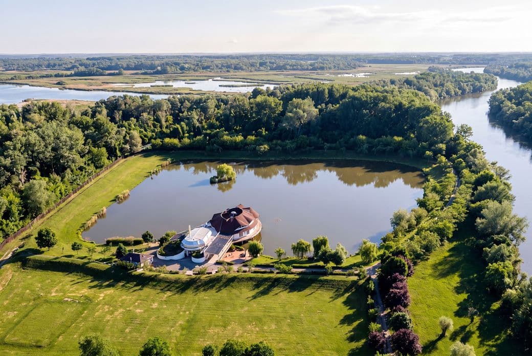 Aerial view of a luxurious house with a circular lake, surrounded by lush greenery and adjacent to a river under a clear sky.