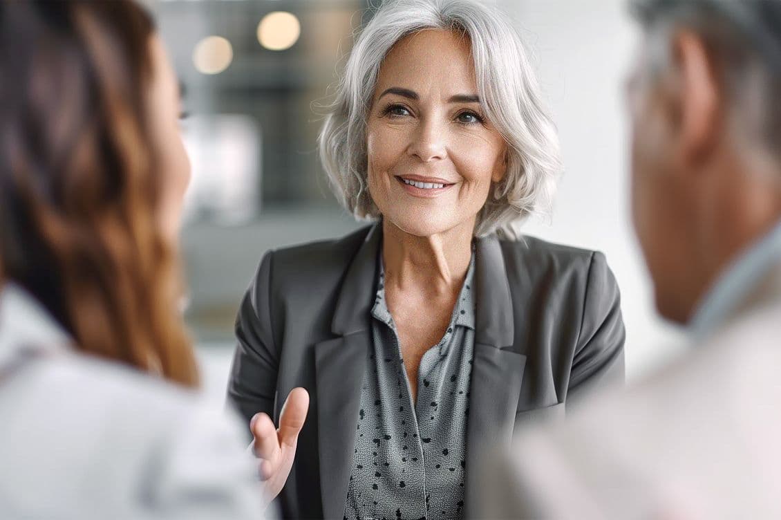 A smiling, gray-haired woman in a gray blazer speaks to two people across a table.