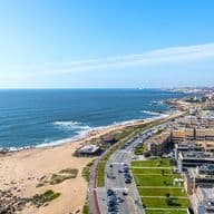 Aerial view of a coastal cityscape with a sandy beach, rocky shoreline, and buildings along a road under a clear blue sky.