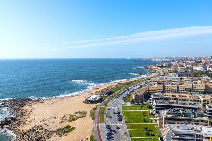 Aerial view of a coastal cityscape with a sandy beach, rocky shoreline, and buildings along a road under a clear blue sky.