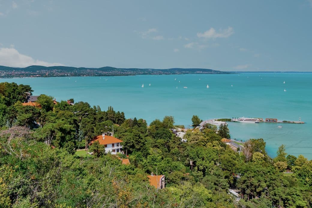 Scenic view of a turquoise lake with sailboats, surrounded by green trees and red-roofed houses, under a clear blue sky.