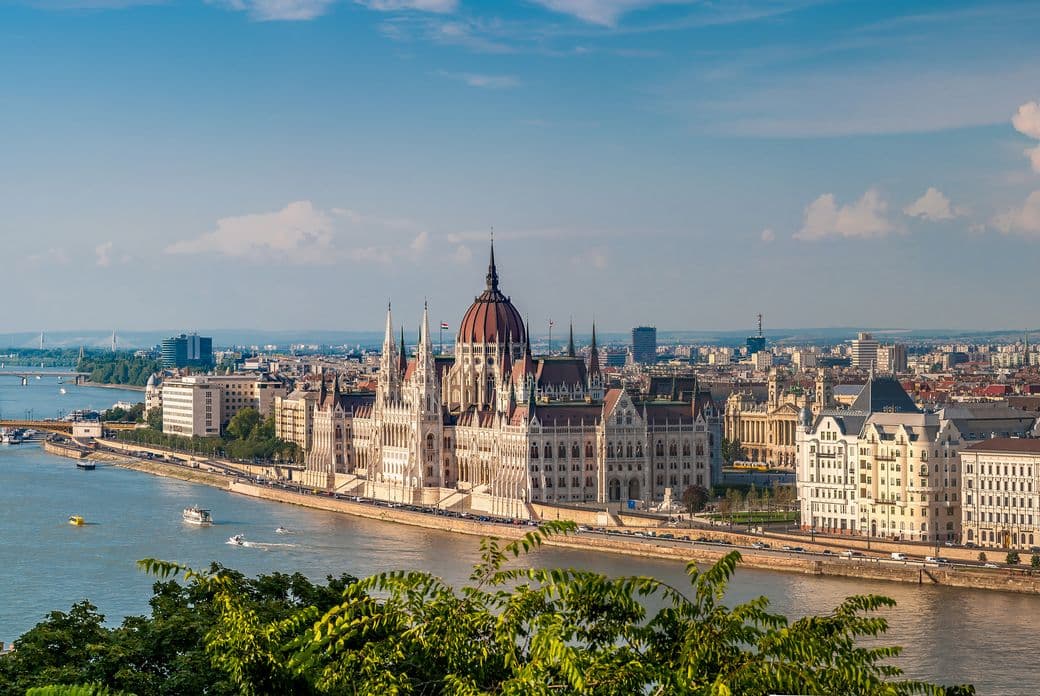 Aerial view of the Hungarian Parliament Building in Budapest, set beside the Danube River under a clear blue sky, with cityscape in the background.