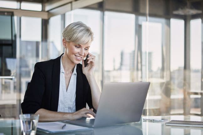 A smiling woman in a black blazer uses a laptop while talking on her cell phone in a bright, modern office.