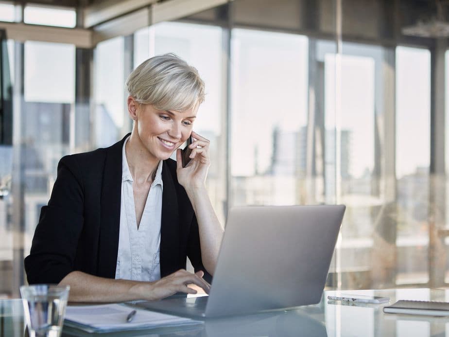 A smiling businesswoman in a modern office, talking on the phone while working on her laptop.
