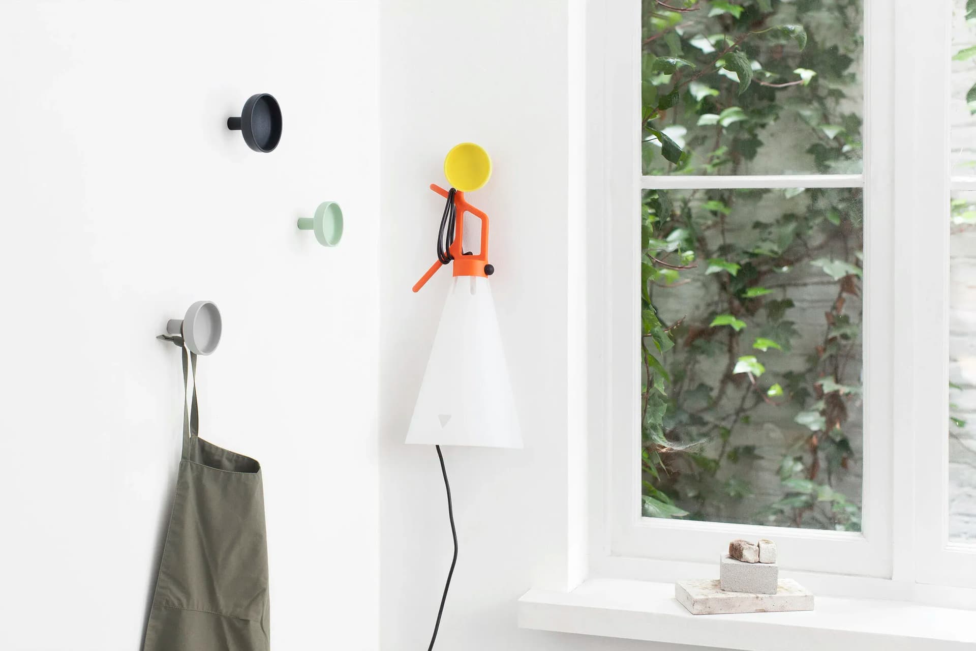 Wall-mounted hooks with a gray apron and a modern cone-shaped lamp beside a window with leafy vines outside.