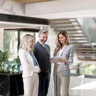 Real estate consultation in a modern luxury property: A female real estate agent in a light gray suit presents property data on a tablet to two interested clients - a woman in a white blazer and a man in a dark suit. The scene takes place in an elegant living space with a floating staircase, marble elements, floor-to-ceiling windows, and natural light. The clients are looking with interest at the presented information.