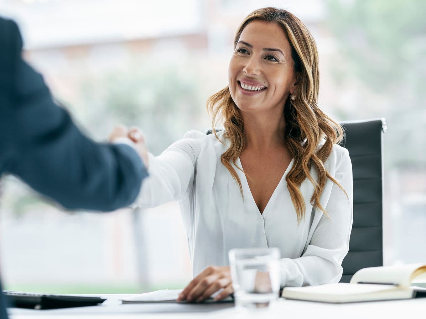 Una donna sorridente in camicetta bianca stringe la mano a un uomo in giacca e cravatta in un ufficio luminoso.