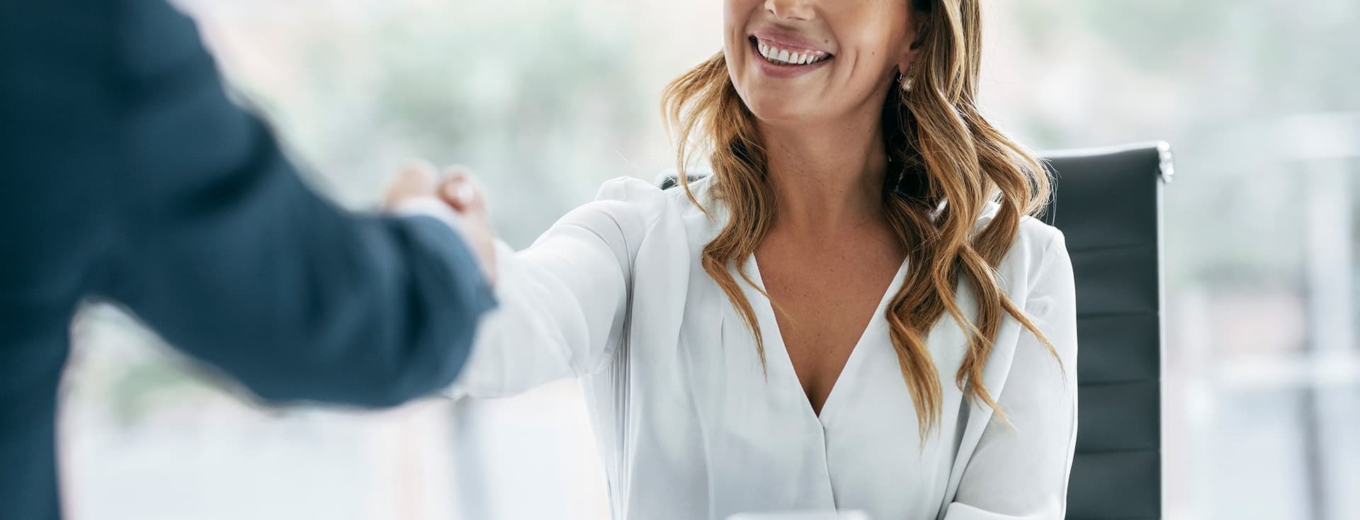 A smiling woman in a white blouse shakes hands with a man in a suit in a bright office.