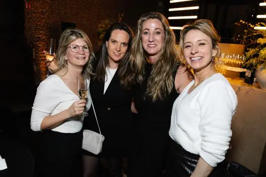 Four women posing at a party, holding champagne glasses, wearing white and black outfits, with a brick wall and bar in the background.