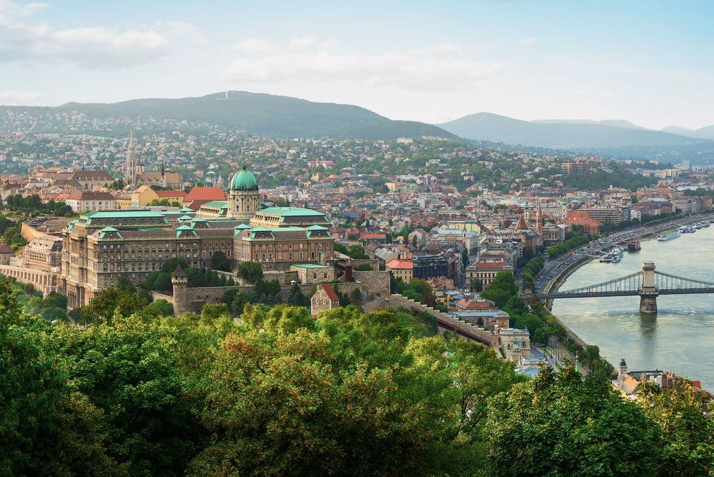 Aerial view of Budapest showing Buda Castle, the Danube River, and Chain Bridge with green hills in the background.