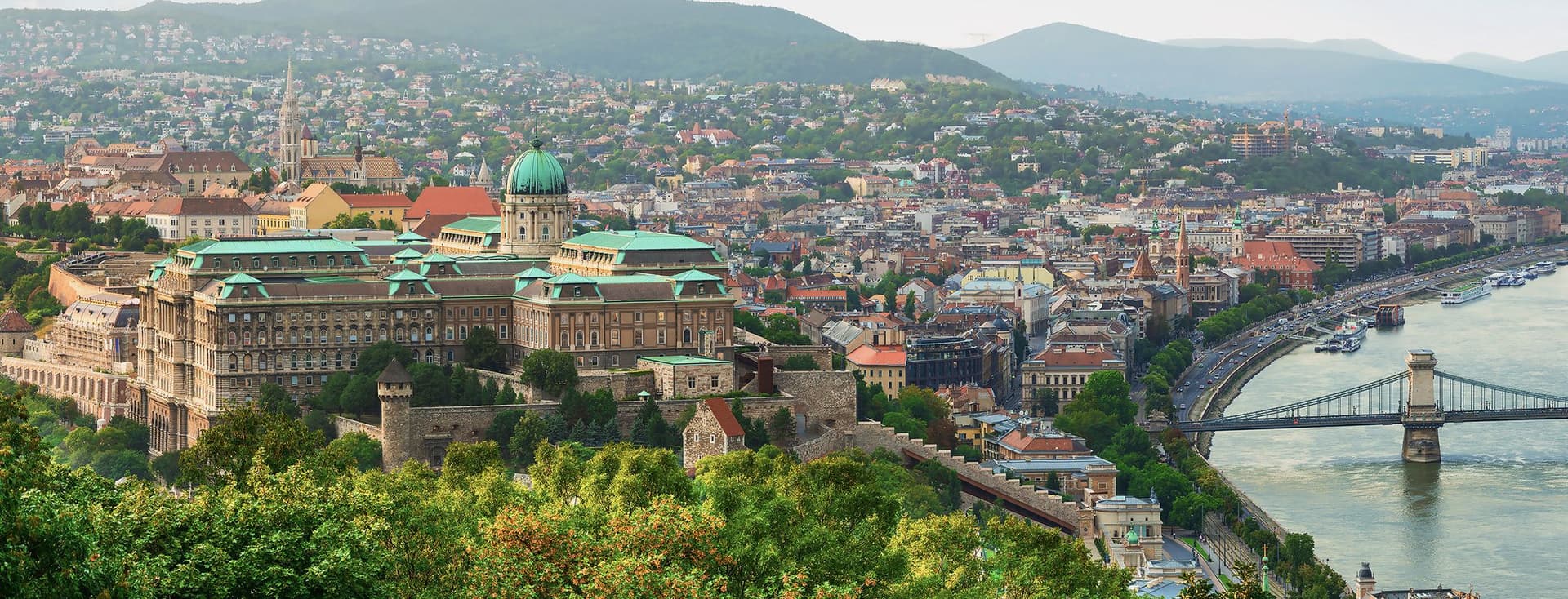Aerial view of Budapest showing Buda Castle, the Danube River, and Chain Bridge with green hills in the background.