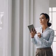 Professional woman in business attire holding a tablet while standing thoughtfully by large windows in a bright, modern office space. She's wearing a white button-up shirt and tailored pants, looking upward in a contemplative pose against a light-filled interior background.