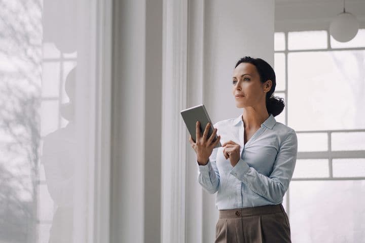 Professional woman in business attire holding a tablet while standing thoughtfully by large windows in a bright, modern office space. She's wearing a white button-up shirt and tailored pants, looking upward in a contemplative pose against a light-filled interior background.