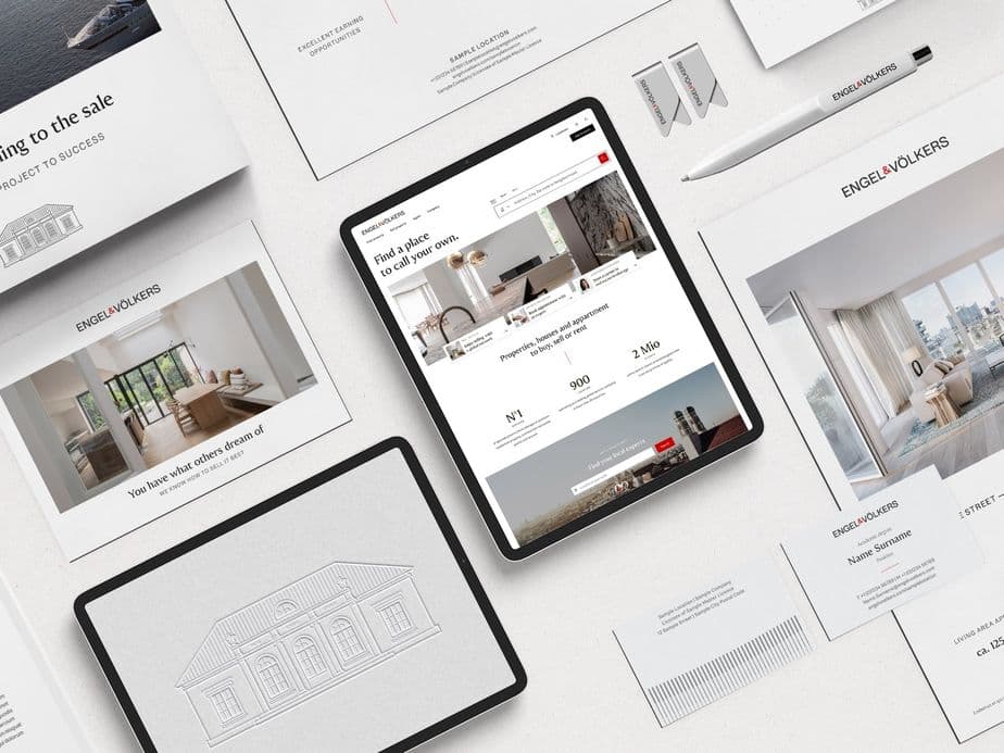 Flat lay photograph of real estate marketing materials including two tablets displaying property listings and architectural drawings, surrounded by various printed marketing collateral with property photos and luxury real estate branding on a white surface. One tablet shows an interactive property listing while the other displays a house sketch.