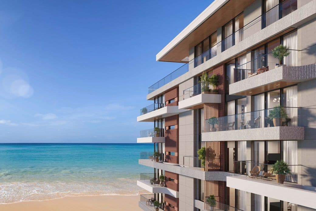 Beachfront residential building with modern architecture, large glass balconies, and greenery, overlooking a calm turquoise sea and sandy shore under a clear blue sky.