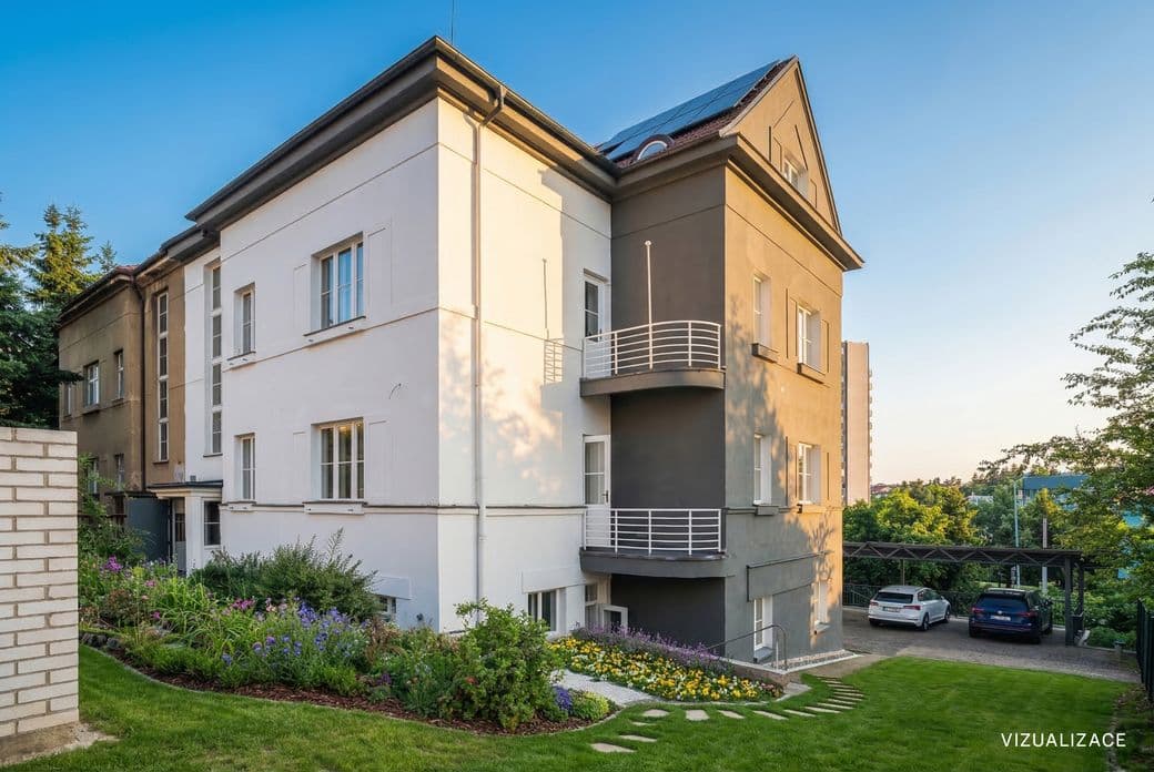View of a corner villa with a light and dark grey façade, two balconies and a pitched roof with windows. In front of the house is a landscaped garden with flowers, lawn and a stone pathway, and on the right there is a covered parking area for cars.