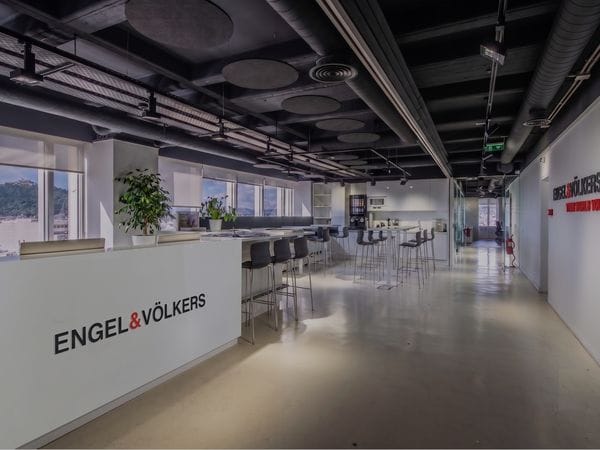 Bright, modern office interior with white counters labeled Engel & Völkers, exposed ceiling ducts, seating area, and large windows with a city view.