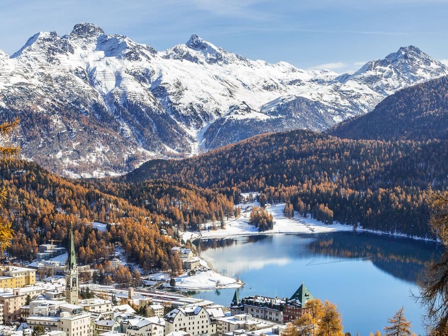 Snow-covered mountains overlooking a serene lake, surrounded by autumn trees and a quaint town with a dusting of snow.