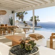 Luxurious open living area of a Mediterranean villa with white walls, wooden beam ceiling and large glass fronts. The space shows a seamless transition between indoor and outdoor areas with views of an infinity pool and the deep blue sea. The furnishings consist of natural materials such as wood and rattan, with a dining table, lounge furniture and a round succulent planting in the foreground. Palm trees and a terrace are visible on the right side.