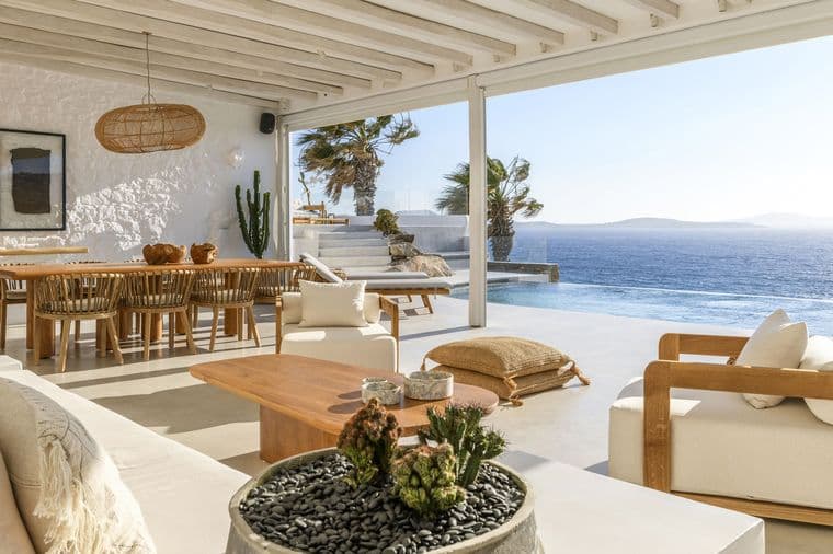 Luxurious open living area of a Mediterranean villa with white walls, wooden beam ceiling and large glass fronts. The space shows a seamless transition between indoor and outdoor areas with views of an infinity pool and the deep blue sea. The furnishings consist of natural materials such as wood and rattan, with a dining table, lounge furniture and a round succulent planting in the foreground. Palm trees and a terrace are visible on the right side.