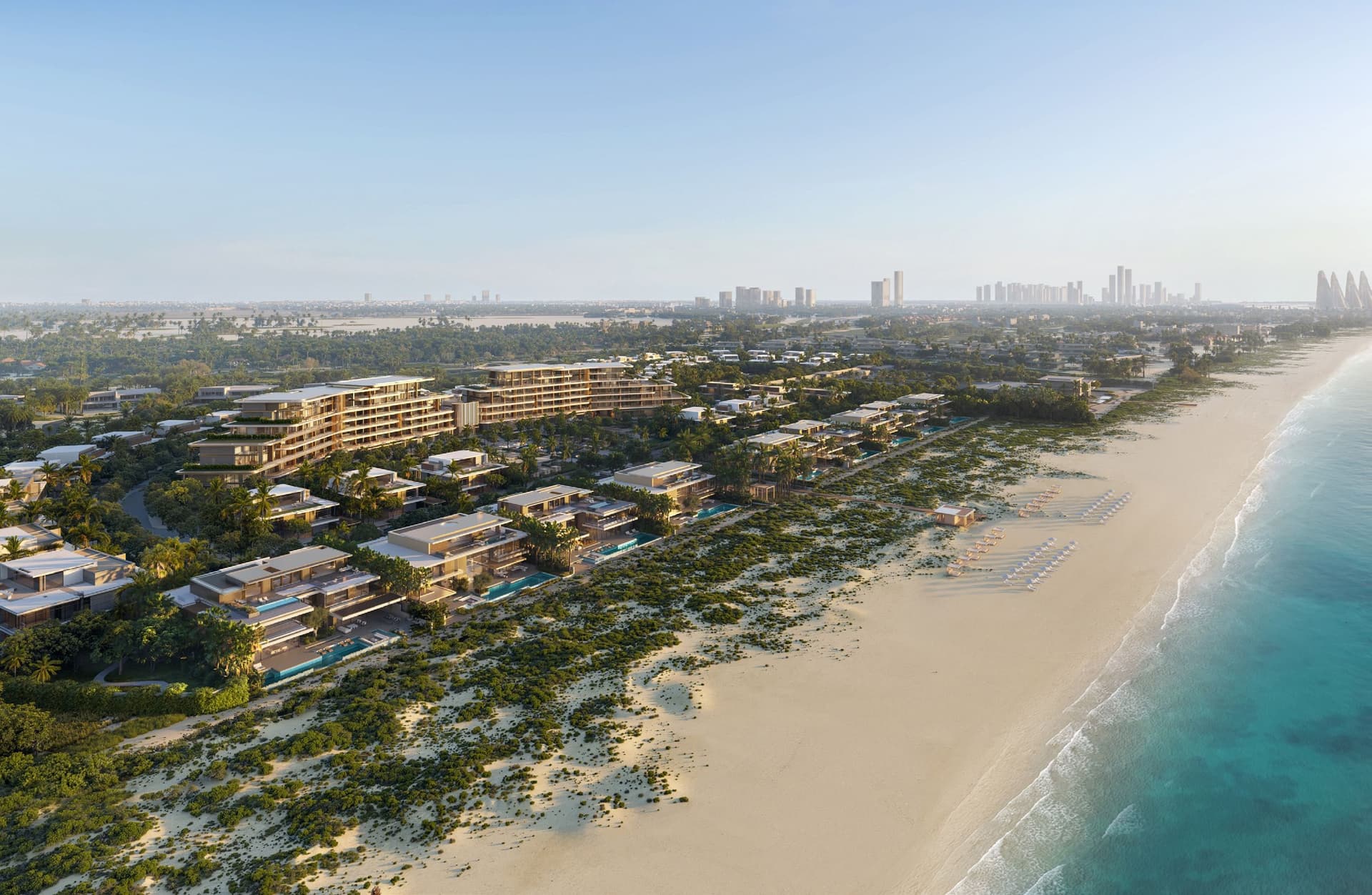 Aerial view of beachfront resort with modern villas, lush greenery, and loungers on a sandy beach beside clear blue water under a bright sky.