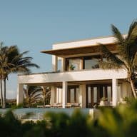 Modern two-story white villa with large windows, palm trees, and an infinity pool, set against a clear blue sky.