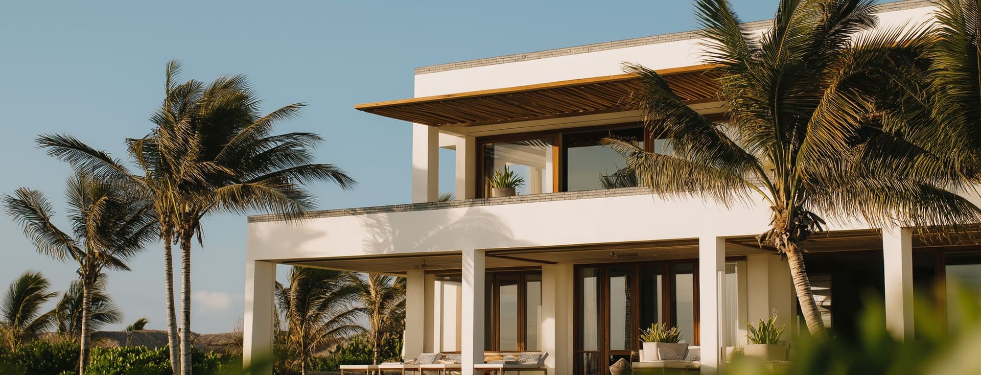 Modern two-story white villa with large windows, palm trees, and an infinity pool, set against a clear blue sky.