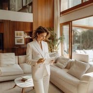 A woman in an elegant white business suit stands in a luxurious living room with wood paneling and large windows, reading documents. The room features cream-colored sofas, natural lighting, and modern interior design elements.