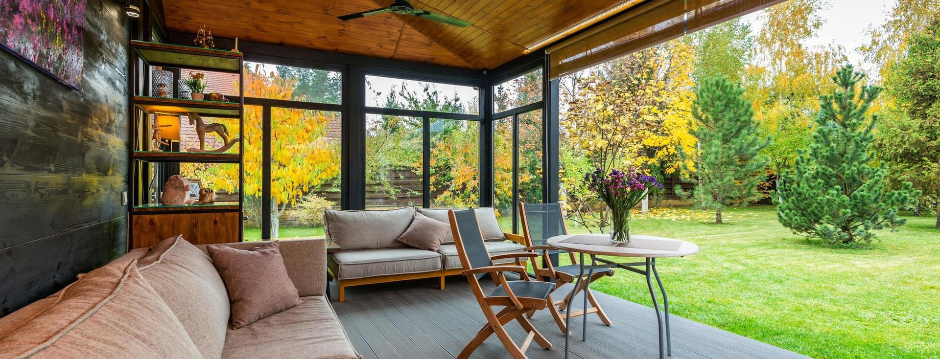 Un porxo càlid i acollidor amb mobles de fusta, una taula i un sofà, amb vista a un jardí ple d'arbres verds i grocs.