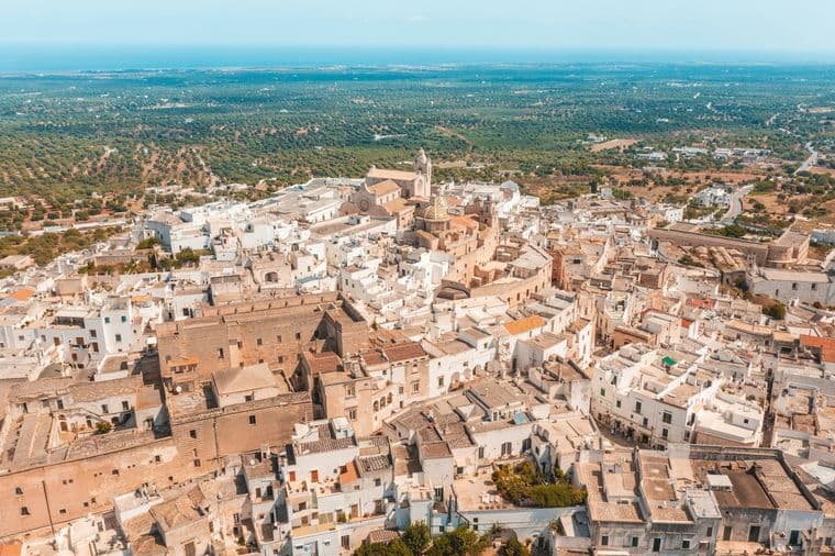 Aerial view of a historic town with white buildings and narrow streets, surrounded by lush greenery under a clear blue sky.