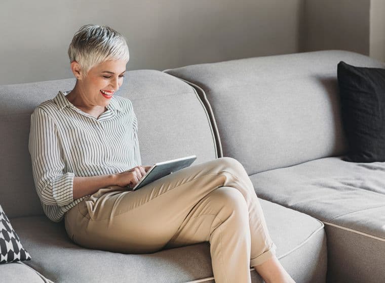 A smiling woman with short white hair sits on a gray sofa, using a tablet. She wears a striped shirt and khaki pants. A black pillow is on the sofa.