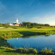 Um sereno campo de golfe com fairways verdejantes, um bunker, um lago refletido e uma igreja branca com campanário ao fundo, sob um céu claro.