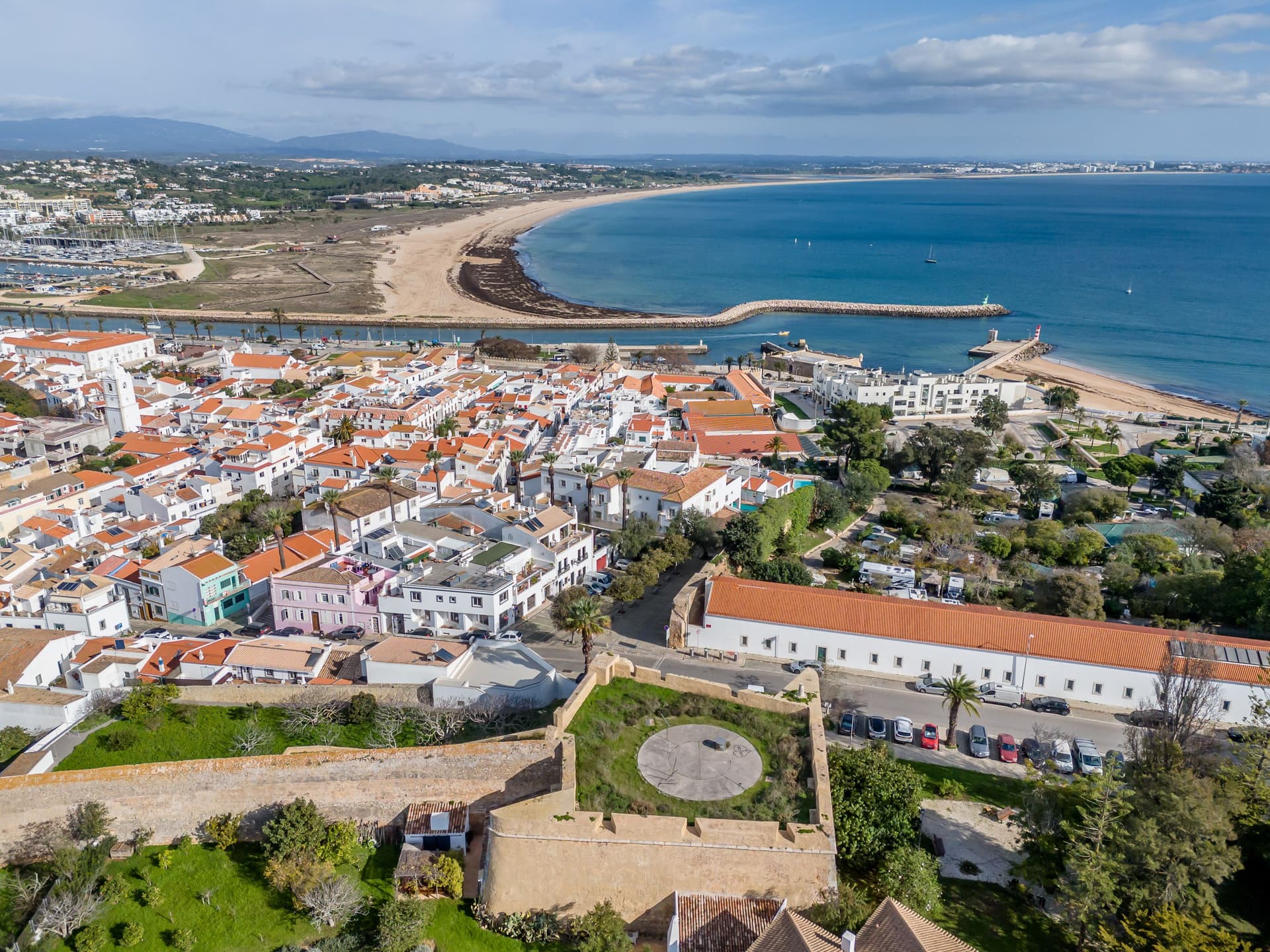 : Vista aérea de Lagos, Portugal, mostrando as muralhas medievais em primeiro plano, o casario histórico de telhados vermelhos, a foz do rio e o mar azul ao fundo