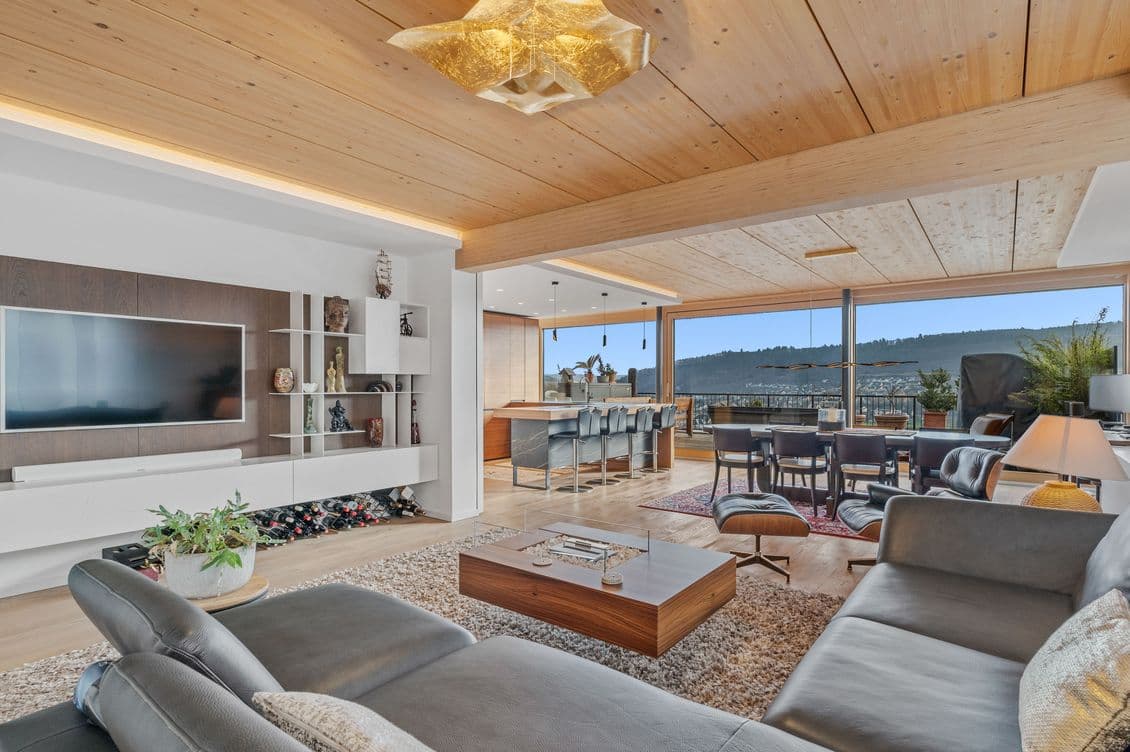 Open living area with light wood paneled ceiling, star-shaped pendant light, gray sofa, white shelves, and large windows overlooking hills.