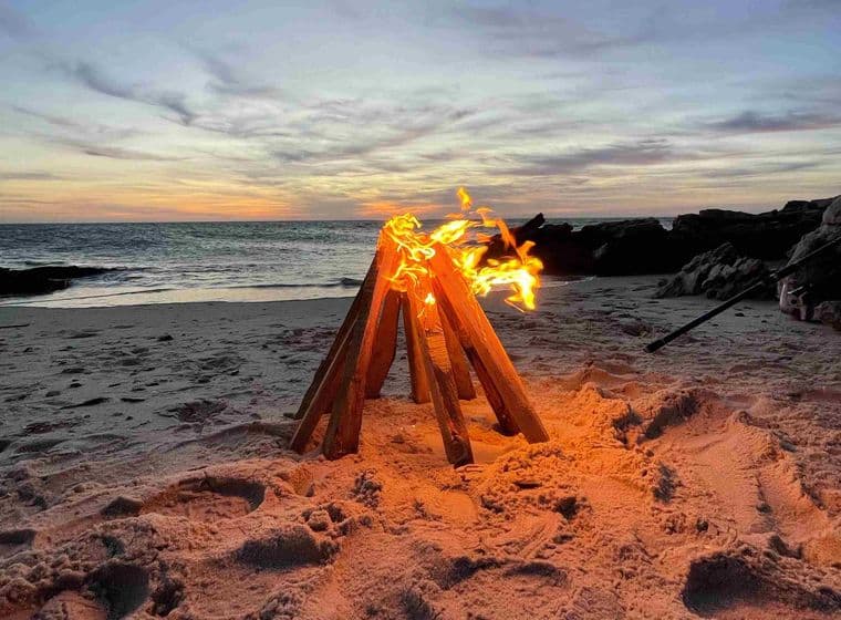 Foguera a la sorra d'una platja al capvespre, amb flames brillants i l'oceà al fons sota un cel amb núvols dispersos.