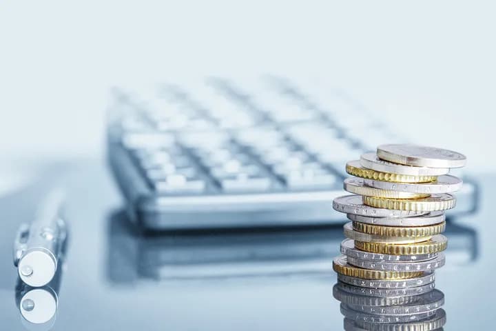 Stack of silver and gold euro coins in focus with a blurred calculator and pen in the background on a light blue surface