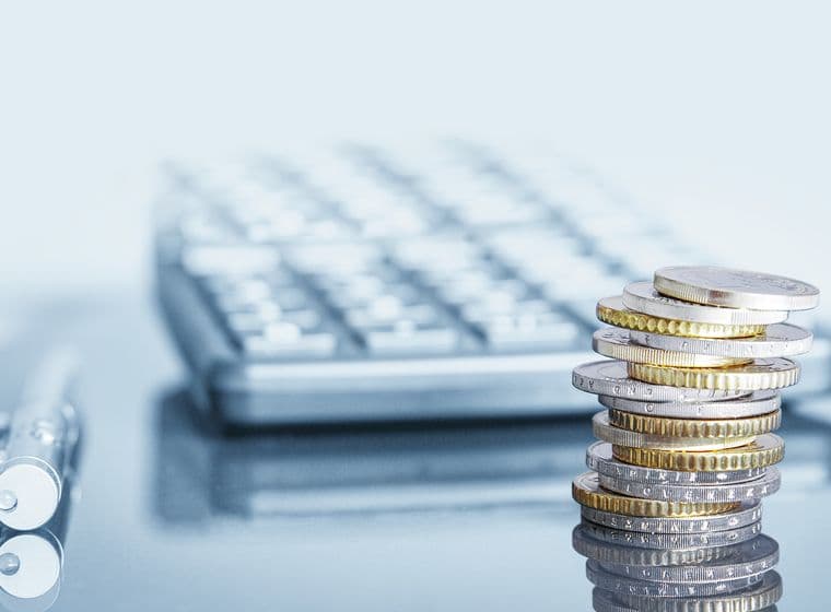Stack of silver and gold euro coins in focus with a blurred calculator and pen in the background on a light blue surface