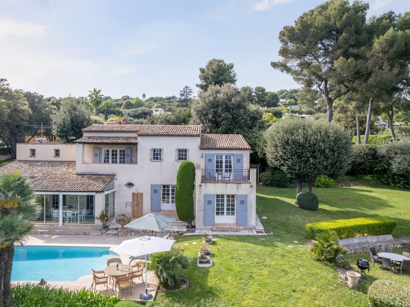 Sunlit villa with blue shutters and terracotta roof, a pool in the foreground, poolside patio, and a lush garden with tall trees.