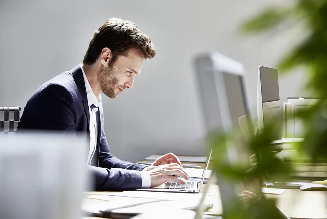 Side profile of a man in a navy blazer typing on a laptop at a desk in a bright office, with monitors and blurred plants in the foreground.