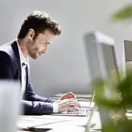 Side profile of a man in a navy blazer typing on a laptop at a desk in a bright office, with monitors and blurred plants in the foreground.