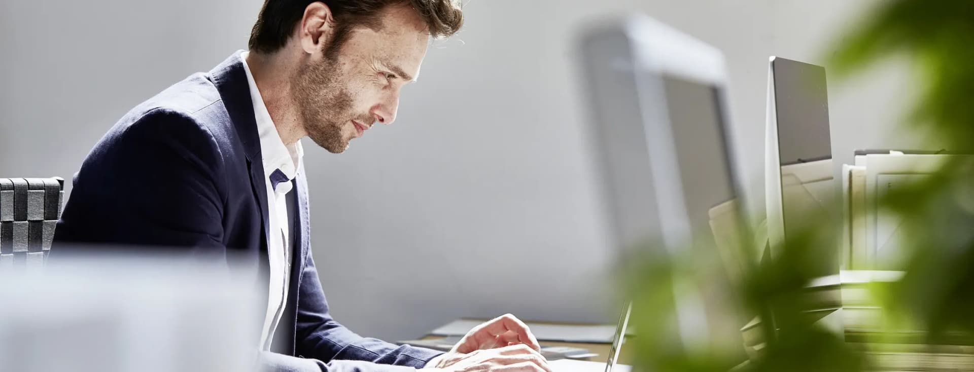 Side profile of a man in a navy blazer typing on a laptop at a desk in a bright office, with monitors and blurred plants in the foreground.