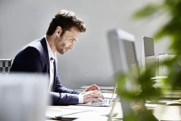 Side profile of a man in a navy blazer typing on a laptop at a desk in a bright office, with monitors and blurred plants in the foreground.