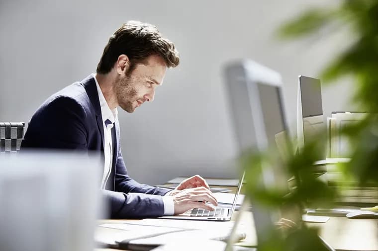 Side profile of a man in a navy blazer typing on a laptop at a desk in a bright office, with monitors and blurred plants in the foreground.