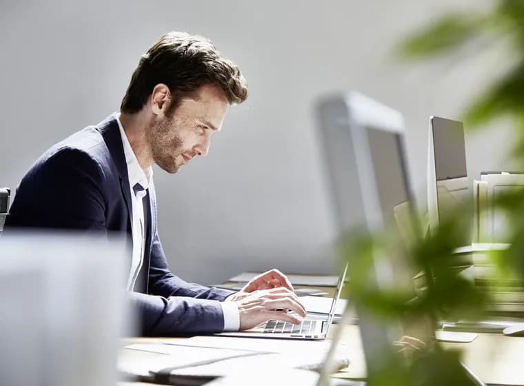Side profile of a man in a navy blazer typing on a laptop at a desk in a bright office, with monitors and blurred plants in the foreground.